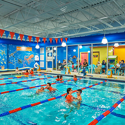 Indoor swimming pool with several people in the water and others seated outside the pool area