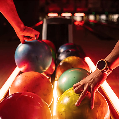 Two hands picking colorful bowling balls from a rack at a bowling alley