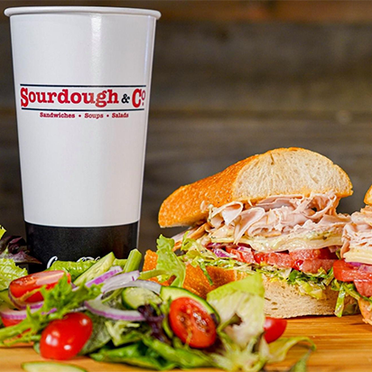 A white cup with the logo 'Sourdough & Co' placed next to a fresh sandwich